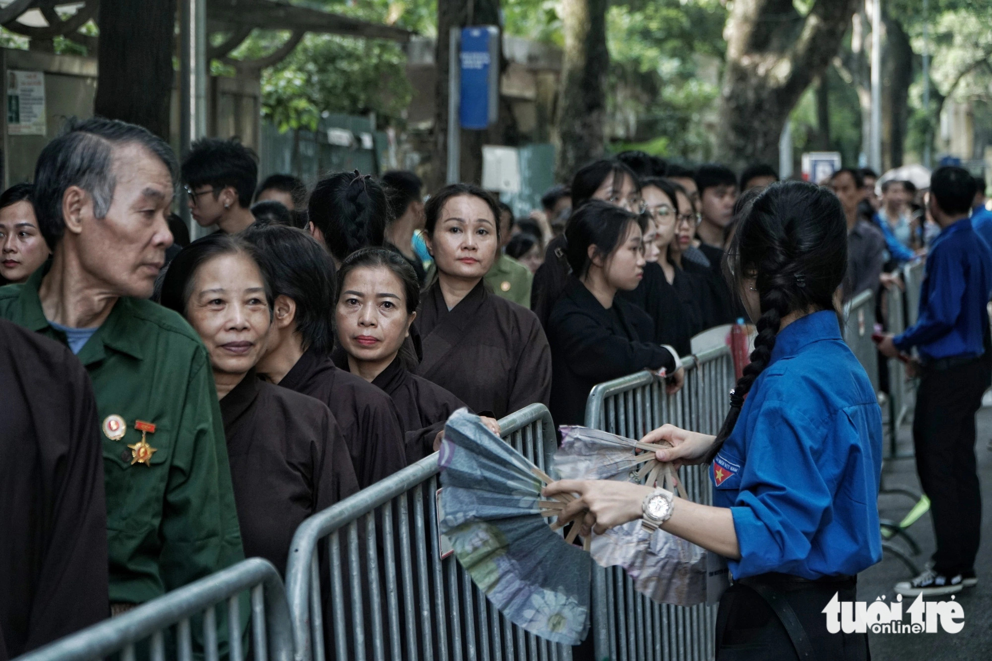 Nguyet viftade med sin solfjäder mot folket: ”Det är väldigt varmt idag, det är många som köar, så vi bestämde oss för att använda solfjädersfläktar för att hjälpa folk att känna sig mindre trötta. Alla kom hit tidigare än oss, från klockan 3 till 4 på morgonen för att ställa sig i kö. Dessutom är det min stolthet att få servera vid generalsekreterare Nguyen Phu Trongs begravning. Idag fick vi mycket tack från folket.” Người dân xếp quạt cây ra đường, pha nước chanh cho người đến viếng Tổng Bí thư Nguyễn Phú Trọng- Ảnh 11.