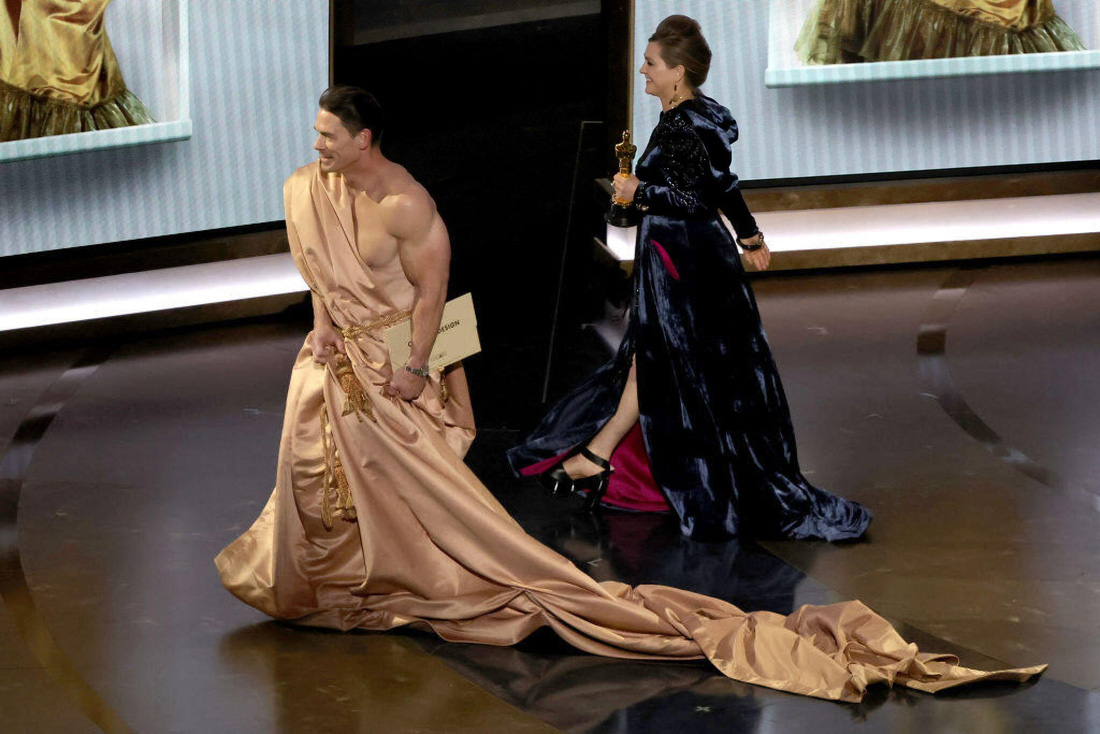 John Cena (trái) &quot;mượn tạm&quot; tấm vải che thân để trở về cánh gà cùng nhà thiết kế thời trang Holly Waddington - Ảnh: GETTY IMAGES