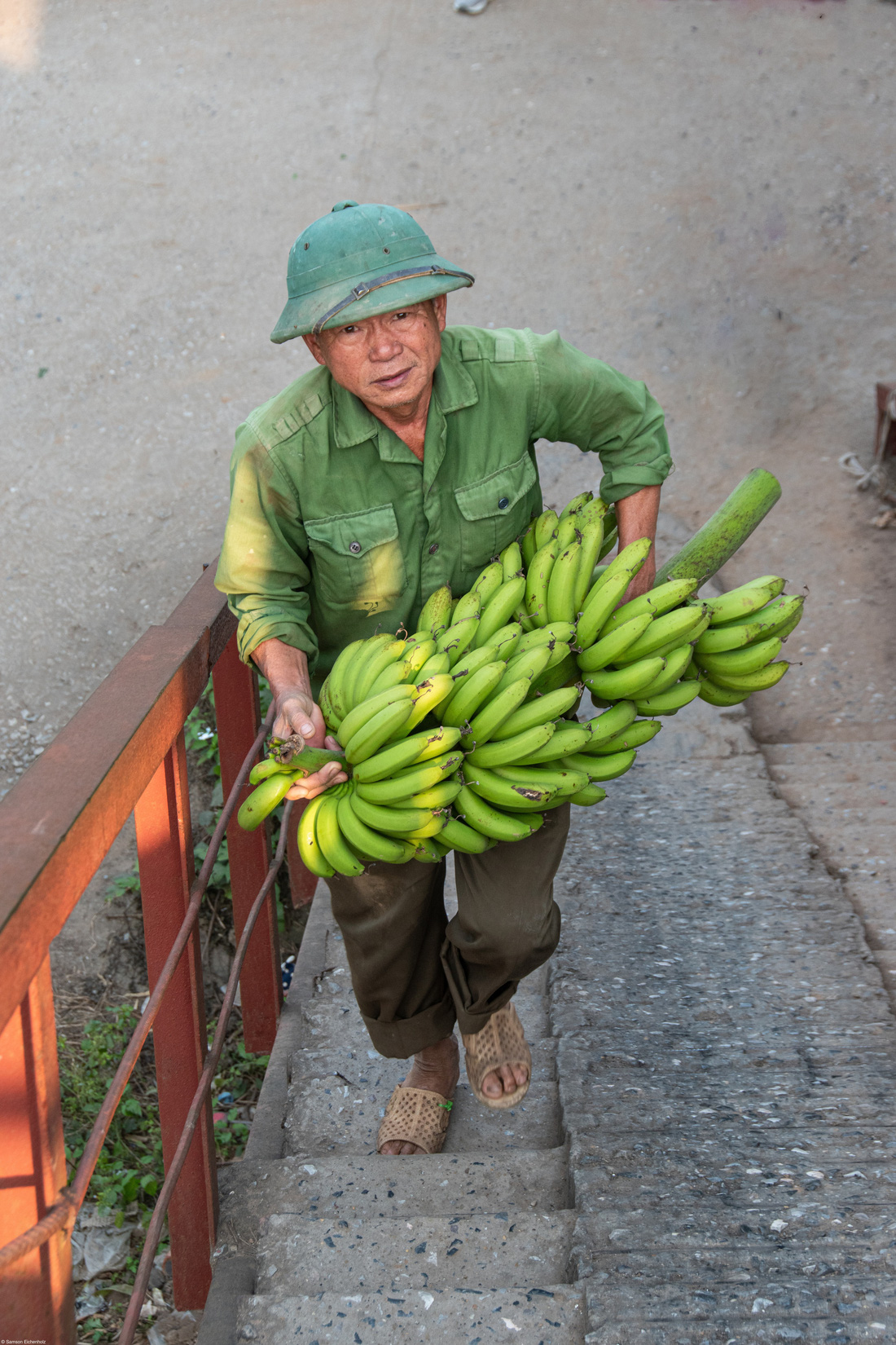 "Dưới cây cầu bắc qua sông Hồng ở Hà Nội, Việt Nam, người dân đã biến đất công thành những khu vườn trù phú. Khi tôi bước xuống cầu thang để khám phá góc khuất này của thành phố, tôi bắt gặp người đàn ông này và ấn tượng với đôi mắt có hồn của ông", tác giả Sam Eichenholz chú thích bức ảnh "Vận chuyển chuối" đoạt giải nhất hạng mục Young 13-17