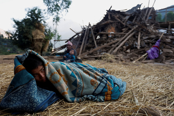 Cậu bé đắp chăn nằm trước ngôi nhà bị đổ sập do động đất ở quận Jajarkot, tỉnh Karnali (Nepal) ngày 5-11. Trận động đất xảy ra ngày 3-11 đã cướp đi sinh mạng của 157 người - Ảnh: REUTERS