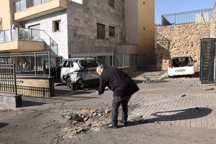 Người dân Israel đứng cạnh một miệng hố và những chiếc xe bị hư hại ở Kiryat Shmona, một trong những thành phố phía bắc Israel bị nã rocket - Ảnh: AFP