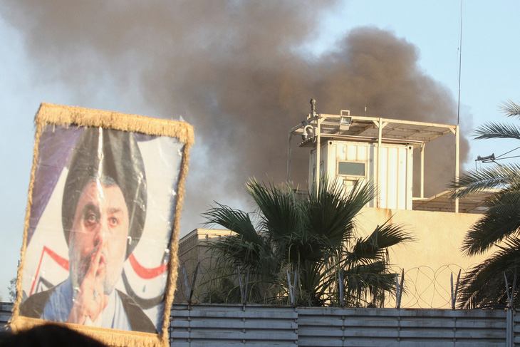 Một người biểu tình giơ cao chân dung giáo sĩ Moqtada al-Sadr khi khói bốc lên từ tòa nhà Đại sứ quán Thụy Điển trong một cuộc biểu tình gần đại sứ quán, vài giờ sau khi họ xông vào và phóng hỏa Đại sứ quán Thụy Điển sáng 20-7 - Ảnh: REUTERS