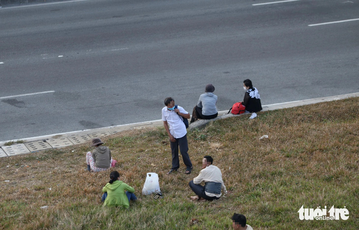 Người dân ngồi trên bãi cỏ ven quốc lộ 1 chờ xe khách đến đón - Ảnh: MINH HÒA