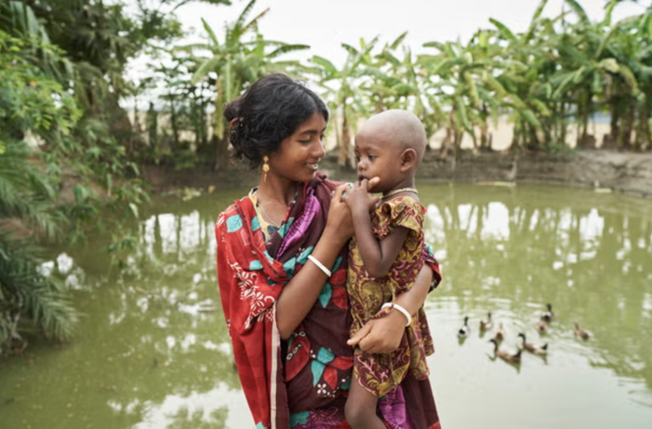 Chị Jhorna (22 tuổi) ẵm con gái Obonita (3 tuổi) đứng trước nguồn nước họ sử dụng hằng ngày - Ảnh: WATERAID