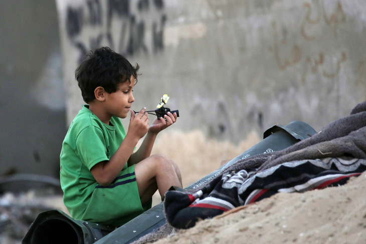 Một đứa bé người Palestine tự tạo niềm vui bên cạnh các thùng đạn rỗng ở TP Khan Yunis, phía nam Dải Gaza, giữa lúc giao tranh Israel - Hamas căng thẳng. (AFP)