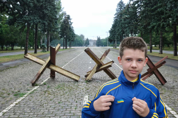 Một cậu bé người Ukraine khoe bộ đồ thể thao mới của đội tuyển quốc gia - món quà em được mẹ tặng trước mùa giải Euro 2024. Phía sau là nhím chống tăng được thiết lập để cản đường xe tăng tại các con đường chính xung quanh nhà ga trung tâm thành phố Odessa và khu vực Kulykove Pole - Ảnh: RICHARD MORGAN