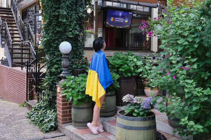 Một cô gái Ukraine đang khoác quốc kỳ lên người và háo hức chờ đợi trận đấu giữa Ukraine và Romania. Trên quốc kỳ, cô viết to cụm từ “Kherson” - tên một thành phố ở Ukraine và cũng là quê hương của cô. Tuy nhiên, cô gái này đã phải ly hương trước các cuộc không kích của Nga - Ảnh: RICHARD MORGAN